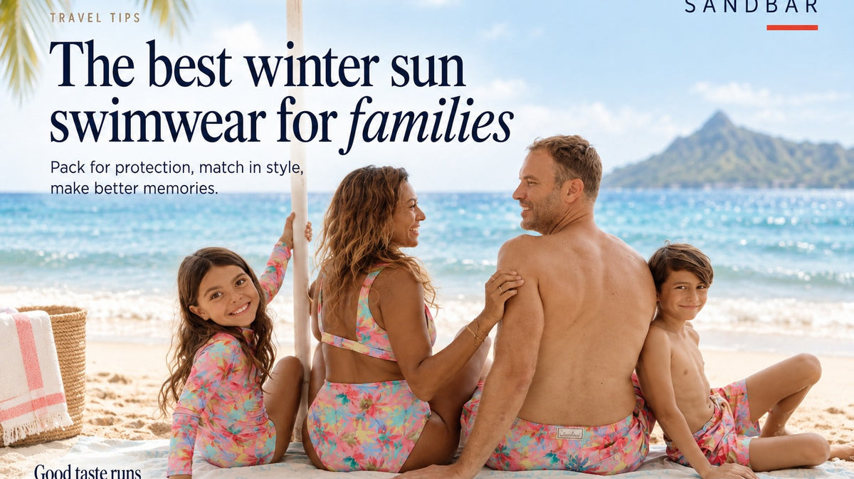 A family of four relaxing on a tropical beach in matching Sandbar Pink Palm swimwear, with turquoise waters and lush mountains behind them. The image features the headline “The best winter sun swimwear for families.