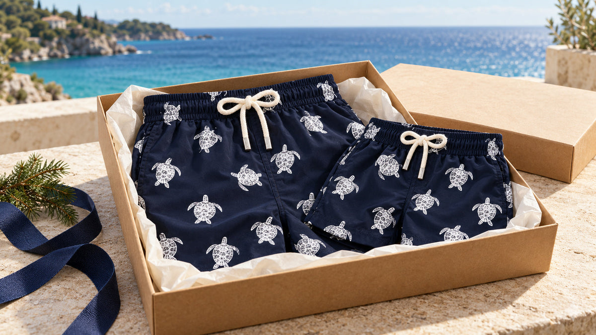 Matching father-and-son turtle swim shorts in a gift box with navy ribbon, overlooking a sunlit blue ocean