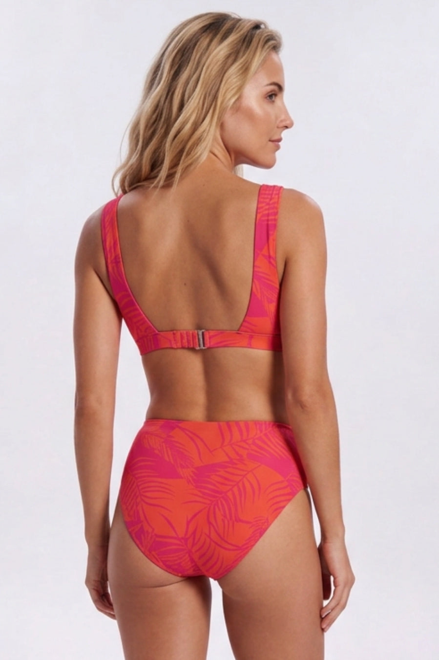A woman shown from the back wearing a two-piece bikini with a bright pink and orange tropical leaf pattern, set against a plain light gray studio background. Sandbar Swimwear