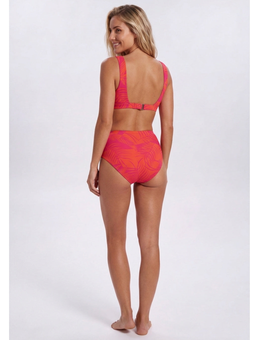A woman modeling a two-piece bikini swimsuit from the back, featuring a bright coral pink and orange tropical leaf pattern, set against a plain light gray studio background. Sandbar Swimwear