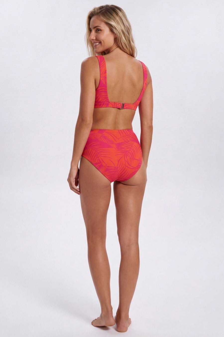 A woman modeling a two-piece bikini swimsuit from the back, featuring a bright coral pink and orange tropical leaf pattern, set against a plain light gray studio background. Sandbar Swimwear