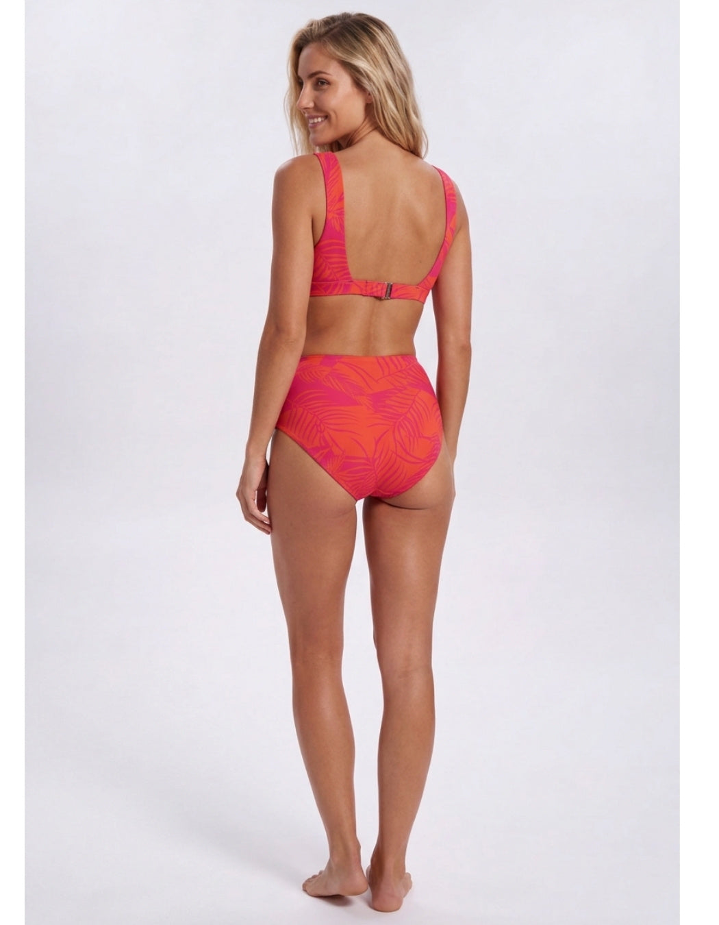 A woman modeling a two-piece bikini swimsuit from the back, featuring a bright coral pink and orange tropical leaf pattern, set against a plain light gray studio background. Sandbar Swimwear