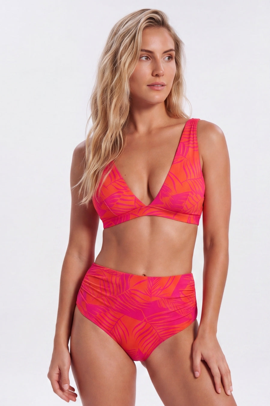 A woman modeling a two-piece bikini with a vibrant pink, orange, and yellow tropical leaf pattern against a plain white studio background. Sandbar Swimwear