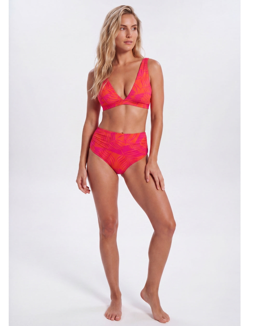 A woman modeling a two-piece bikini swimsuit with a vibrant pink and orange abstract pattern, set against a plain light gray studio background. Sandbar Swimwear