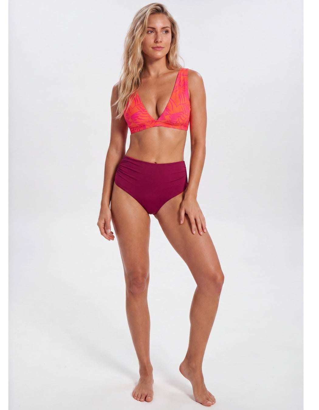 A woman modeling a two-piece bikini set against a plain light gray studio background. The bikini top is bright coral-orange with a subtle tonal pattern, and the high-waisted bottoms are a deep magenta-purple. Sandbar Swimwear