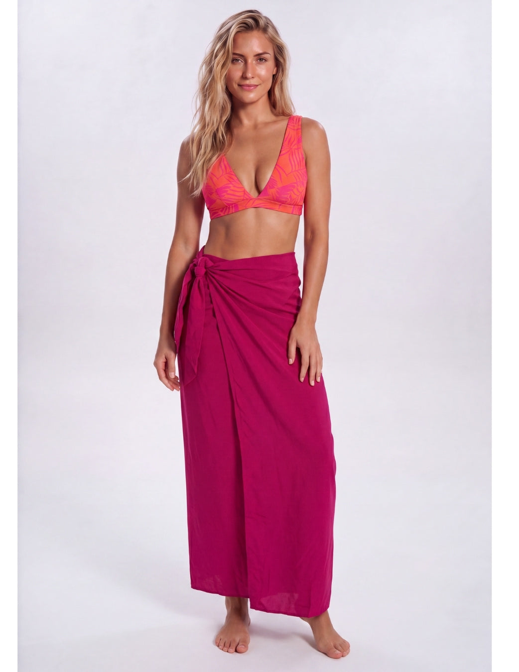 A woman modeling a bright magenta wrap sarong and a matching pink-orange patterned bikini top against a light gray studio background. Sandbar Swimwear
