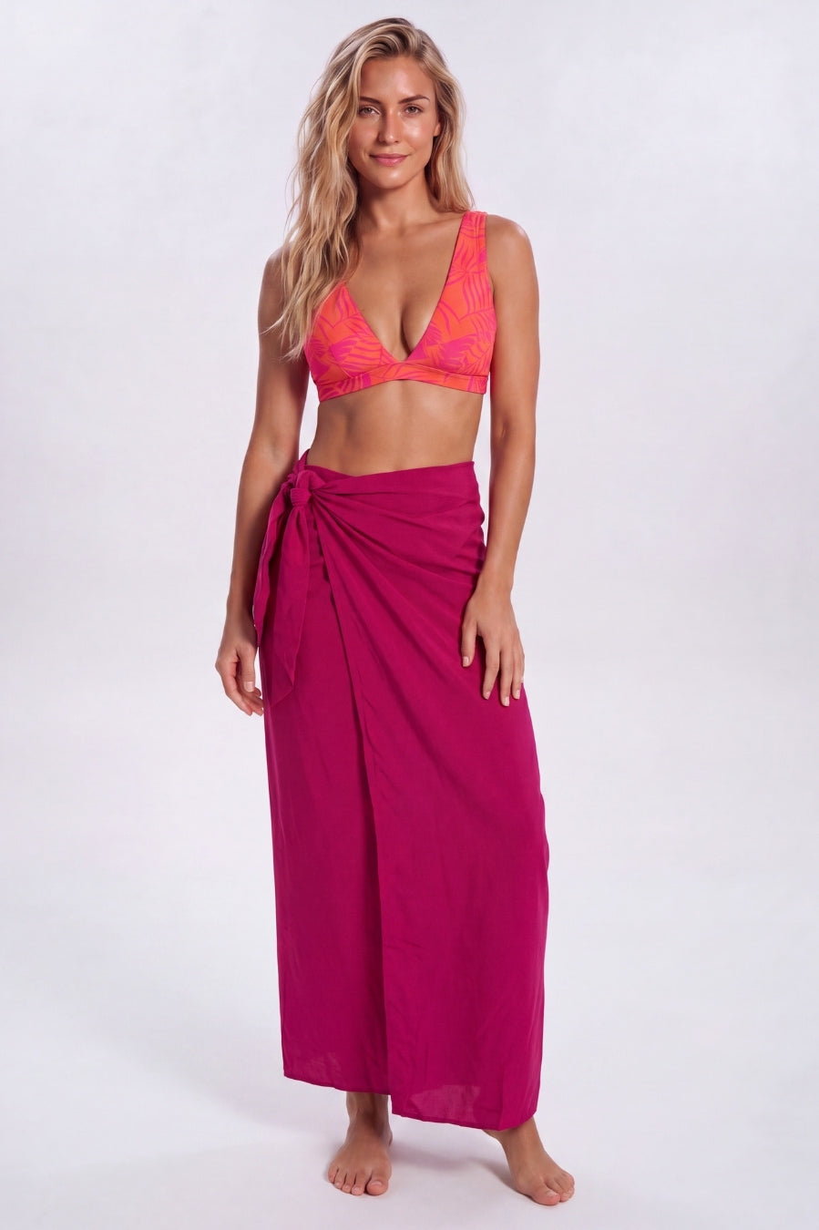 A woman modeling a bright magenta wrap sarong and a matching pink-orange patterned bikini top against a light gray studio background. Sandbar Swimwear