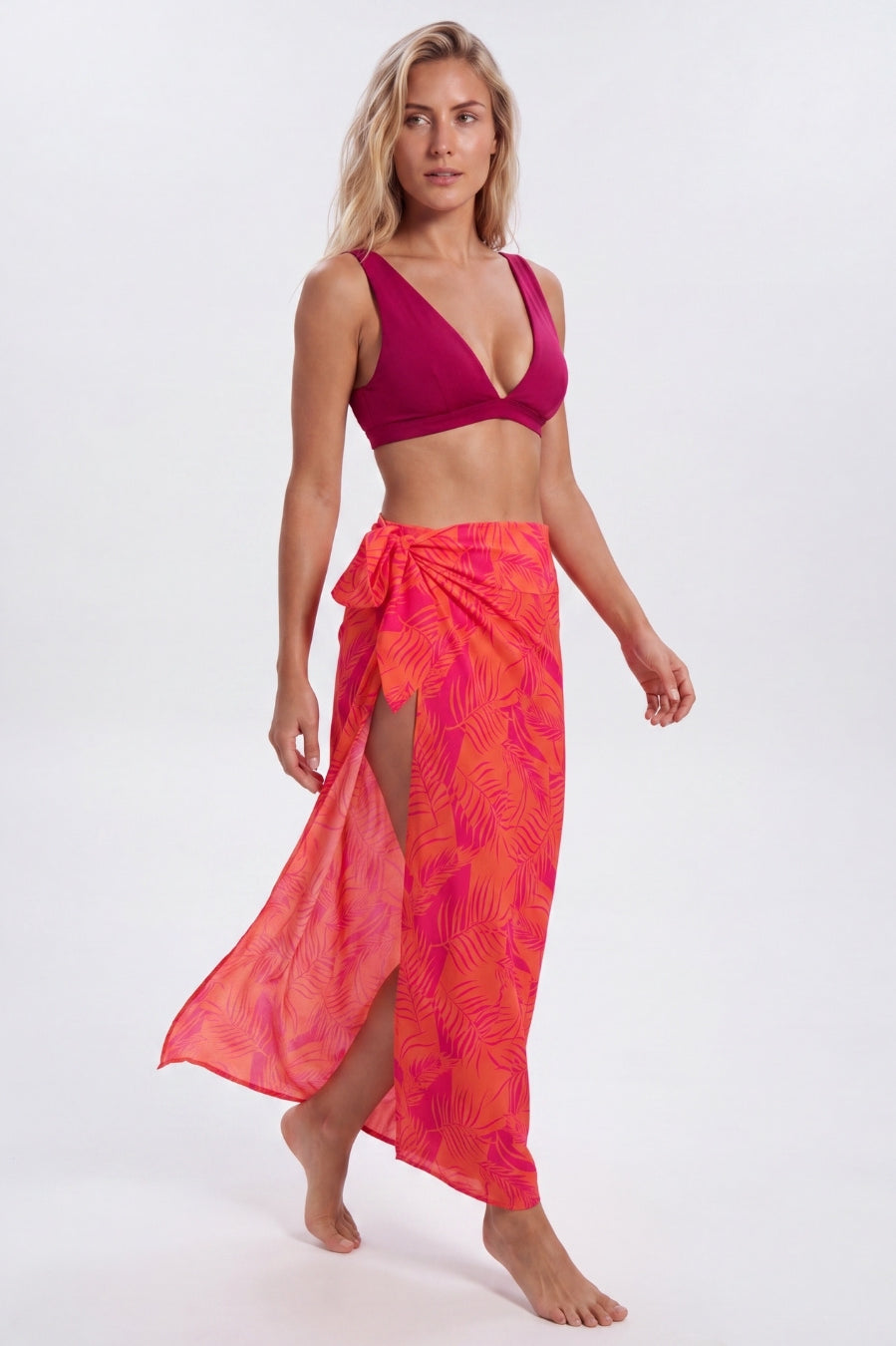 A woman modeling a bright pink and orange tropical leaf-patterned sarong wrap skirt with a matching pink bikini top, standing against a light gray studio background. Sandbar Swimwear