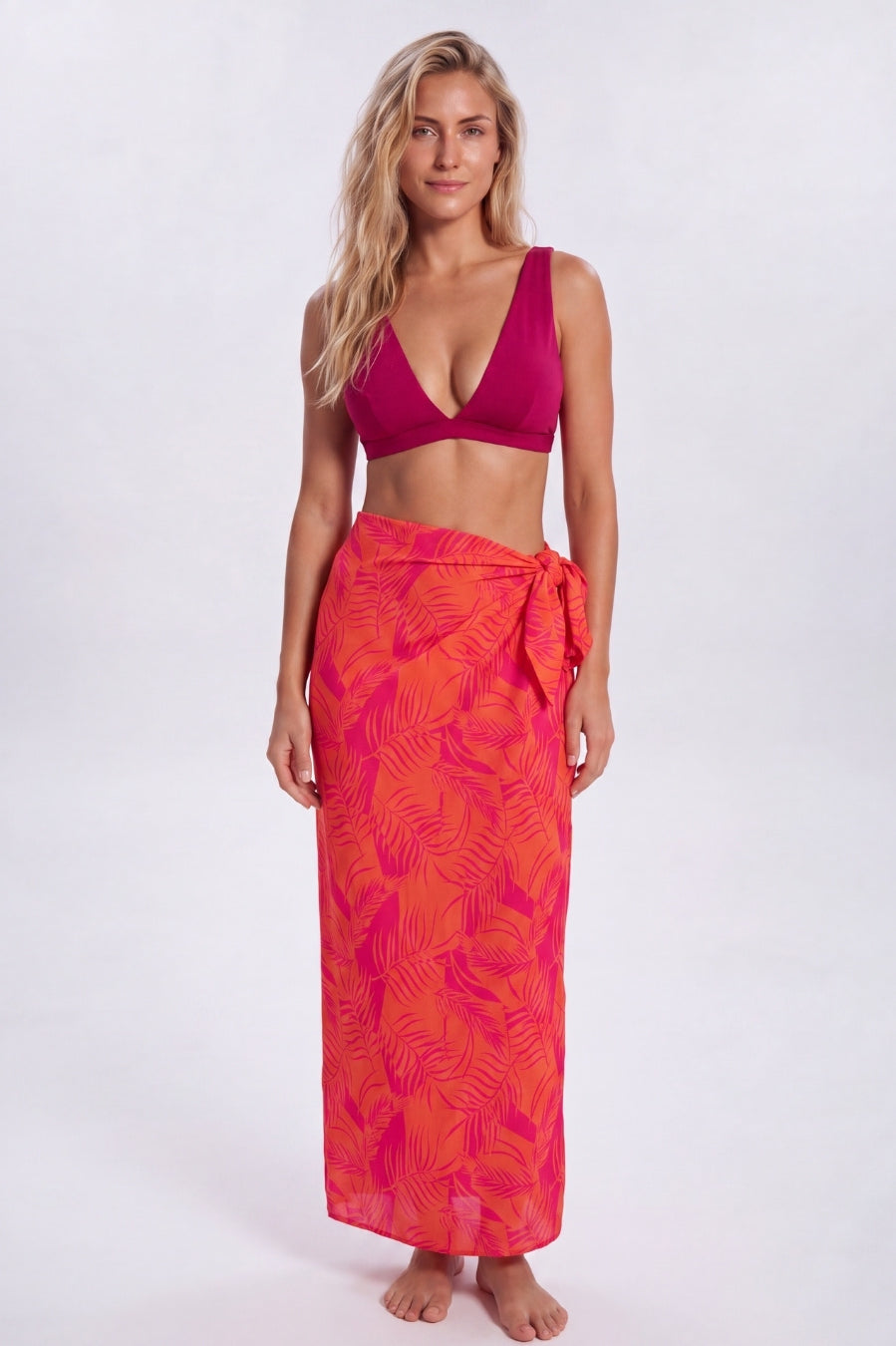 A woman modeling a two-piece swimwear set consisting of a deep magenta pink bikini top and a matching orange and pink tropical leaf print sarong wrap skirt, shot against a plain light gray studio background. Sandbar Swimwear