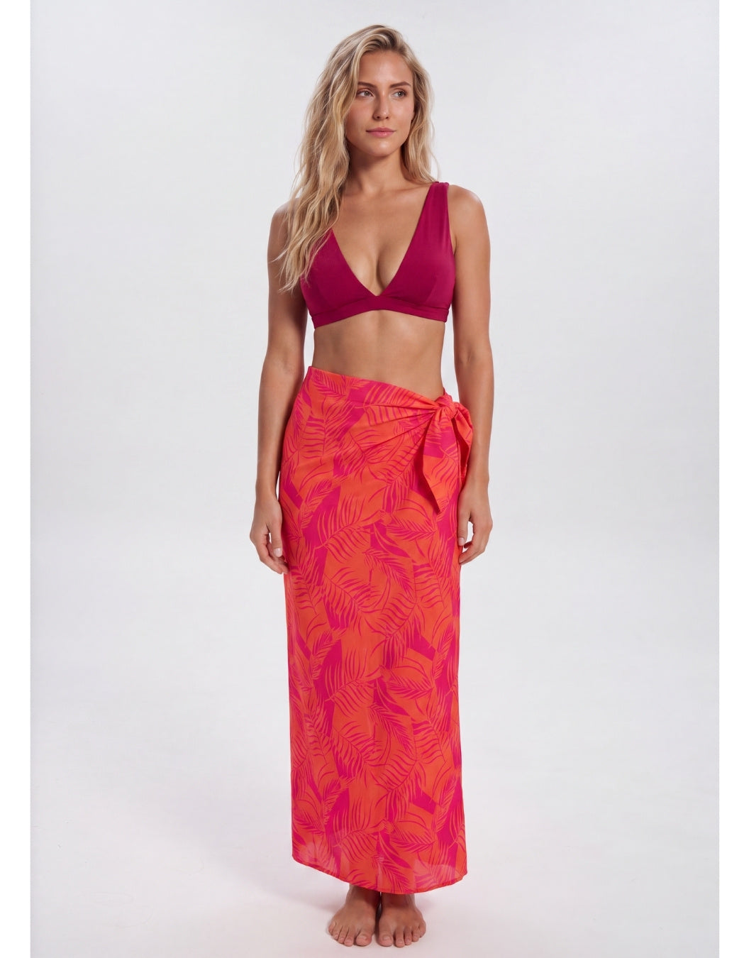 A woman modeling a two-piece swimwear set consisting of a deep magenta-burgundy bikini top and a bright pink-orange tropical leaf print sarong wrap skirt, shot against a seamless light gray studio background. Sandbar Swimwear