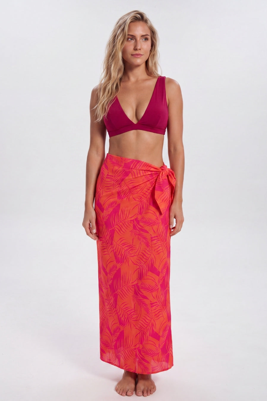 A woman modeling a two-piece swimwear set consisting of a deep magenta-burgundy bikini top and a bright pink-orange tropical leaf print sarong wrap skirt, shot against a seamless light gray studio background. Sandbar Swimwear