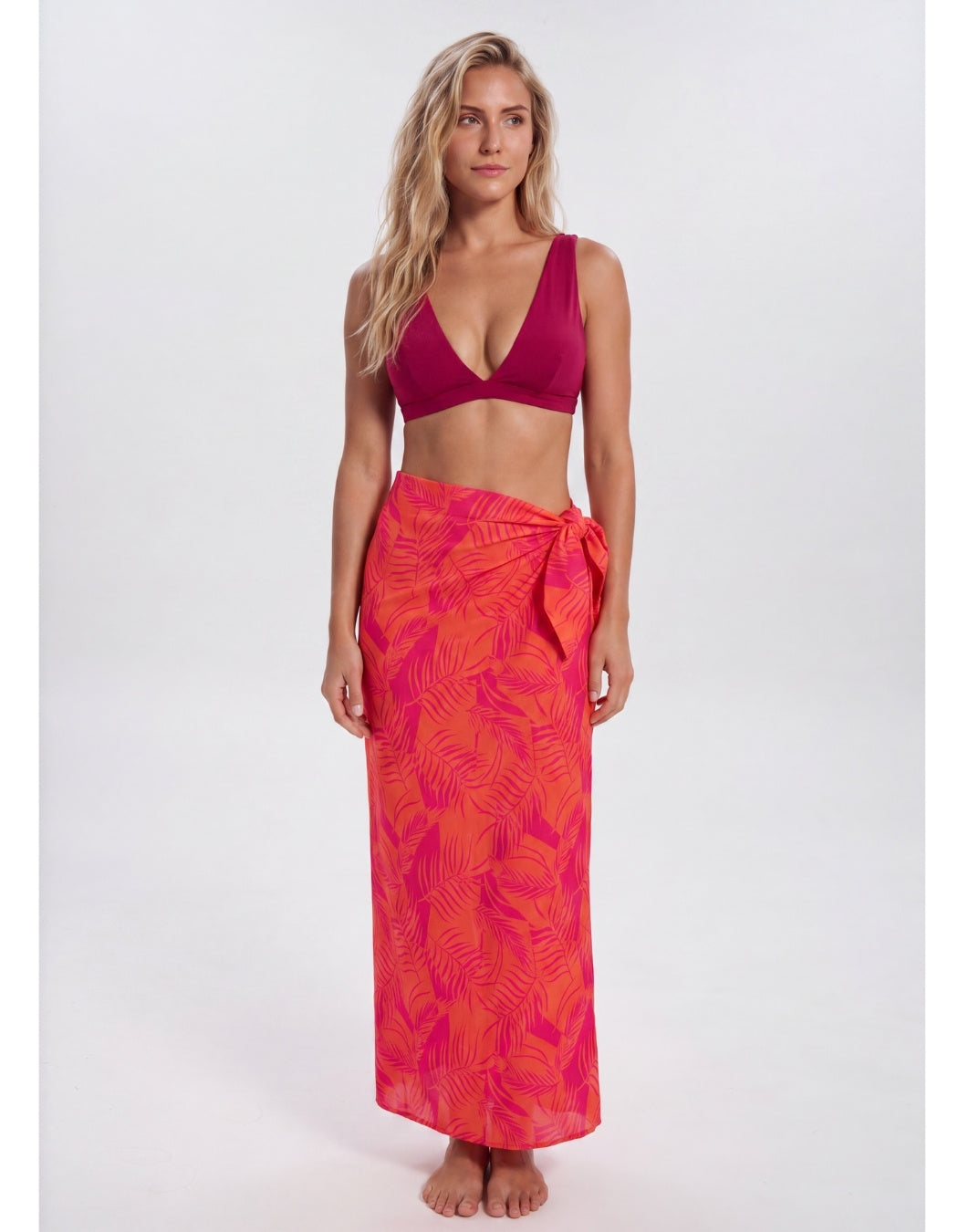 A woman modeling a two-piece swimwear set consisting of a deep magenta-burgundy bikini top and a bright pink-orange tropical leaf print sarong wrap skirt, shot against a seamless light gray studio background. Sandbar Swimwear