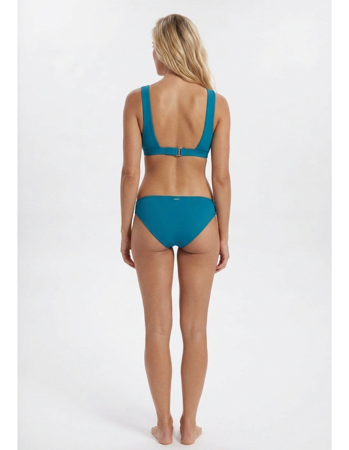A woman viewed from the back wearing a teal two-piece bikini against a plain light gray studio background Sandbar Sustainable Swimwear