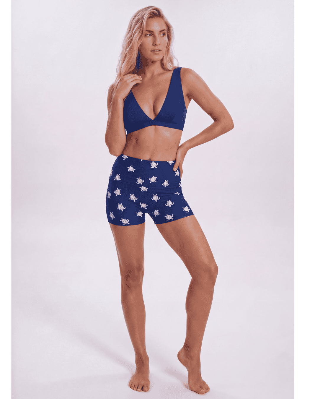A woman modeling a matching two-piece swimwear set consisting of a navy blue V-neck bikini top and high-waisted shorts with a white star pattern, set against a plain light gray studio background. Sandbar Swimwear