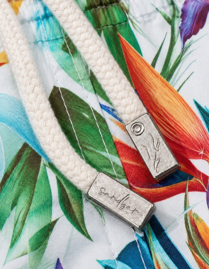 Close-up of a white braided drawstring with a silver metal toggle engraved with the brand name 'Sandbar', resting on a white fabric background with a colorful tropical floral print. Sandbar Swimwear shorts detail
