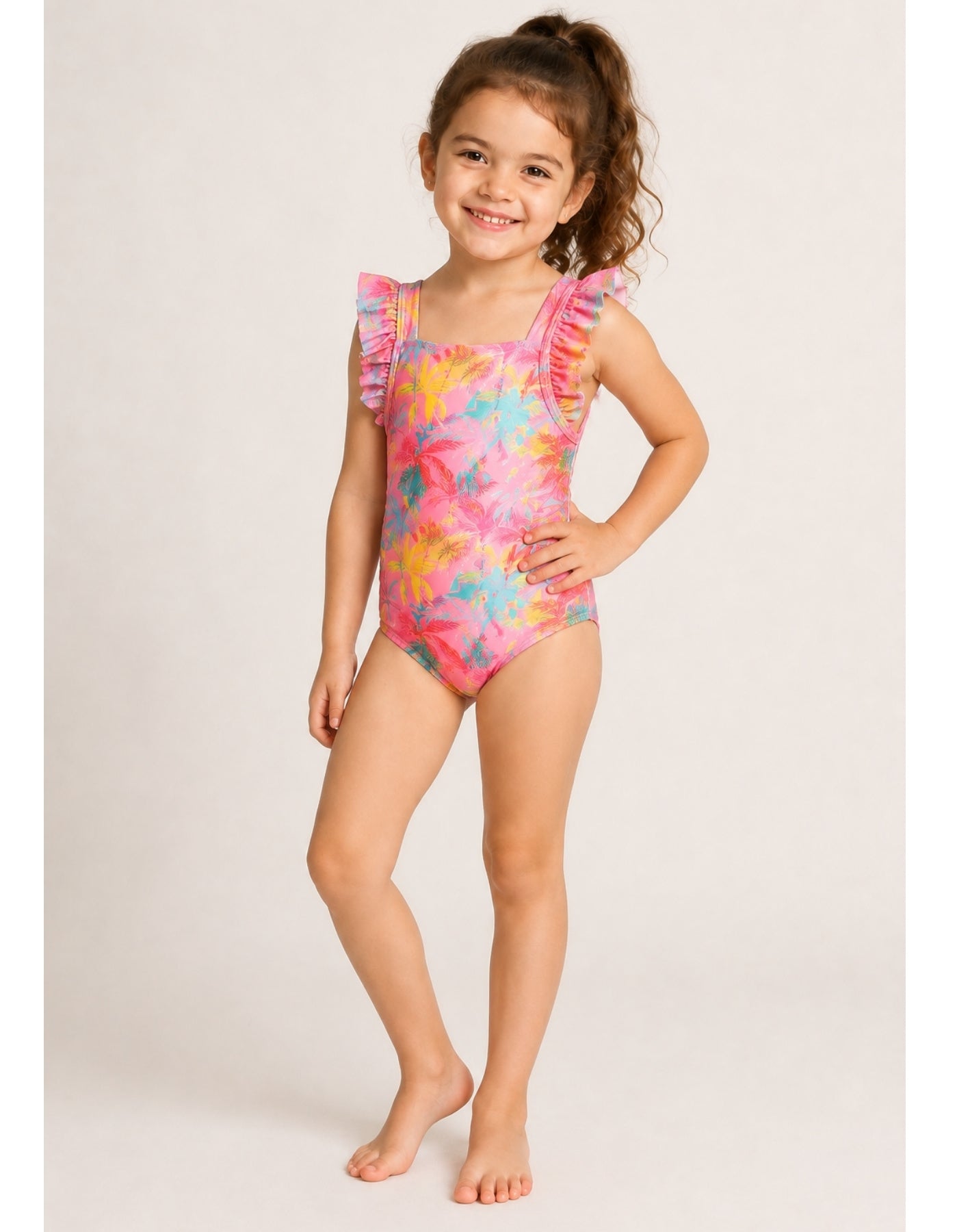 A young girl with curly brown hair stands barefoot against a light beige studio background, wearing a one-piece swimsuit with a bright pink base and a multicolor tropical floral print in yellow, blue, and orange. The swimsuit features ruffled shoulder straps.