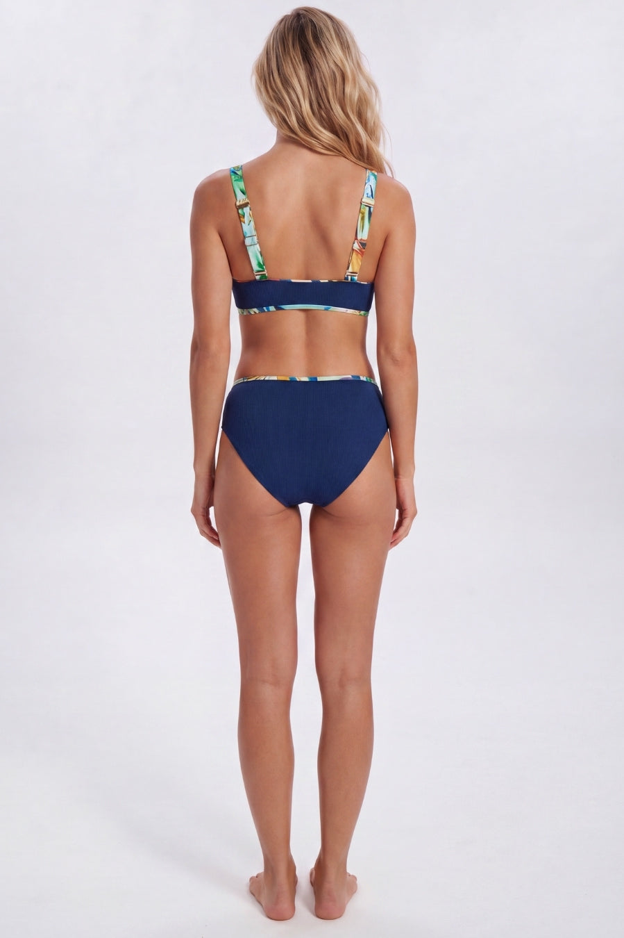 A woman shown from the back wearing a two-piece bikini. The bottom is solid navy blue, while the top has a tropical leaf print with green, yellow, and blue on a white base. Sandbar Swimwear