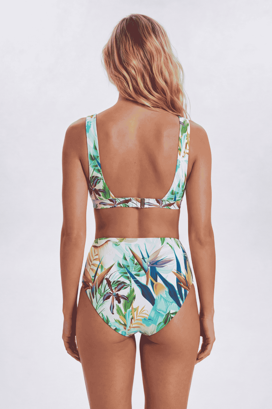 A woman shown from the back wearing a two-piece bikini with a tropical floral print. The swimsuit features a light blue, green, and white pattern with brown and orange accents, set against a plain light gray studio background. Sandbar Swimwear