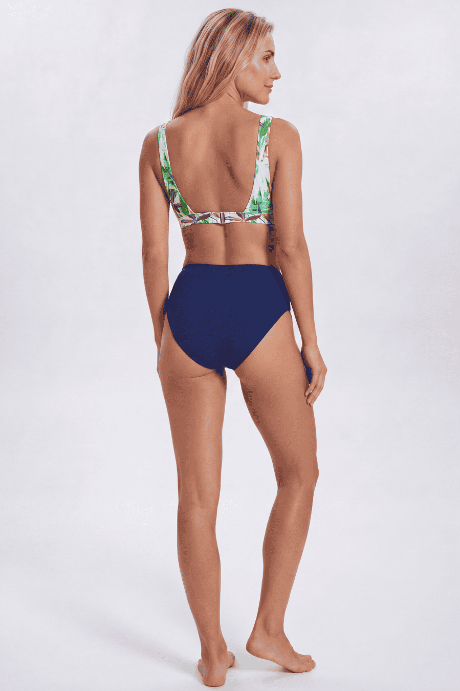 A woman modeling a two-piece bikini from the back, featuring a solid navy blue high-waisted bottom and a green tropical leaf print halter top, against a light gray studio background. Sandbar Swimwear