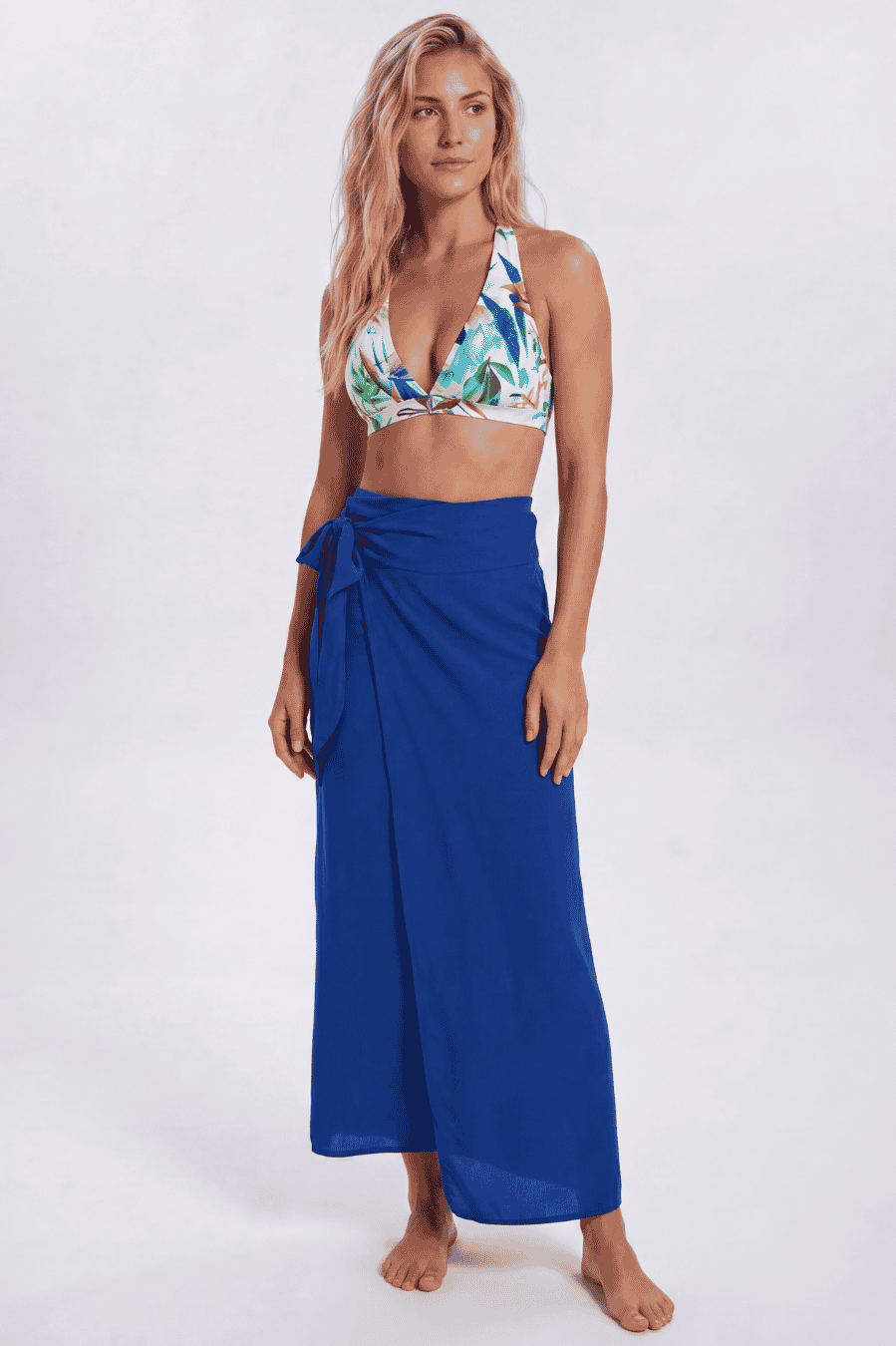 A woman modeling a royal blue wrap sarong skirt paired with a tropical print bikini top, standing against a light gray studio background. Sandbar Swimwear