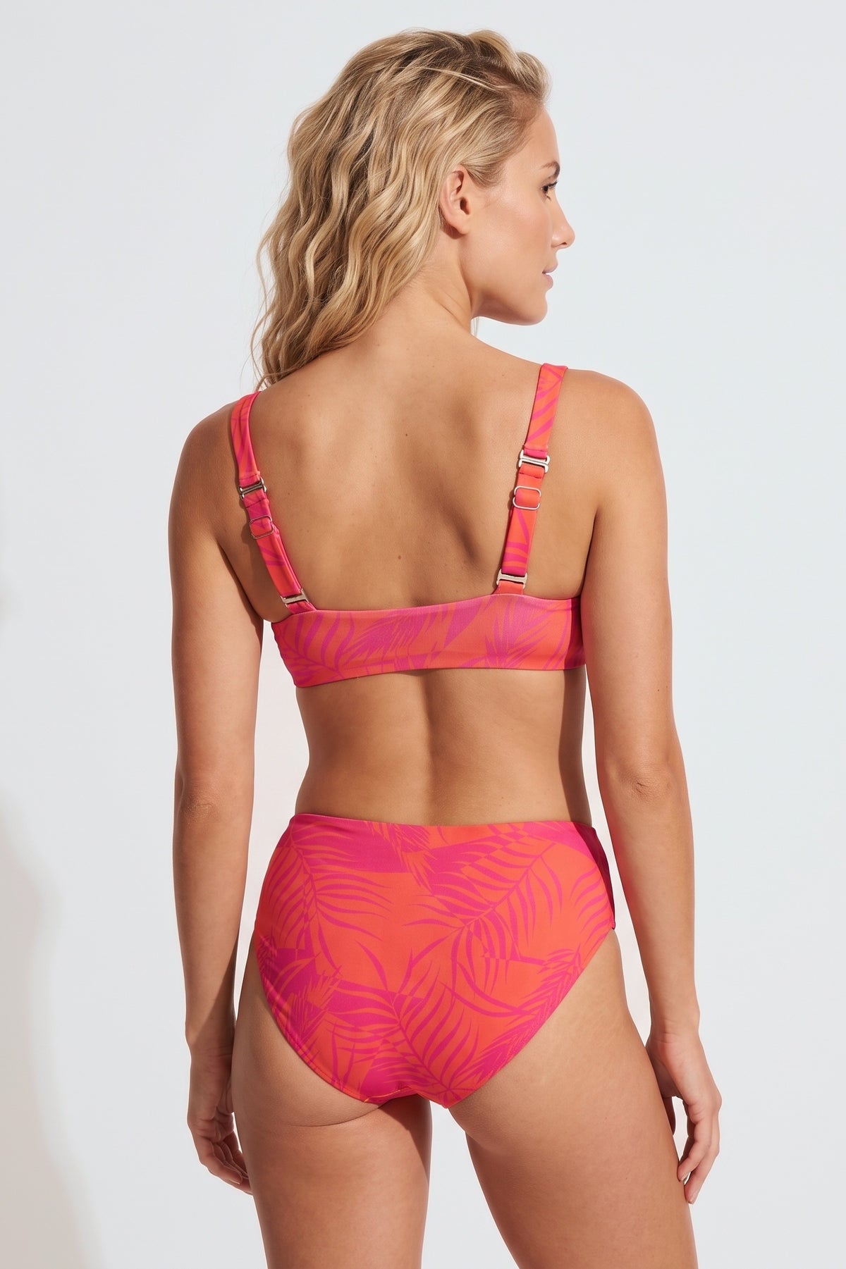 A woman modeling a two-piece bikini from the back, featuring a vibrant pink and orange tropical leaf pattern, set against a plain light gray studio background.