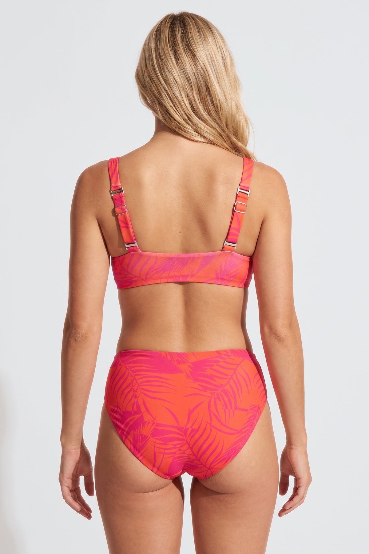 A woman shown from the back wearing a two-piece bikini with a bright pink and orange tropical leaf pattern, set against a plain light gray studio background.