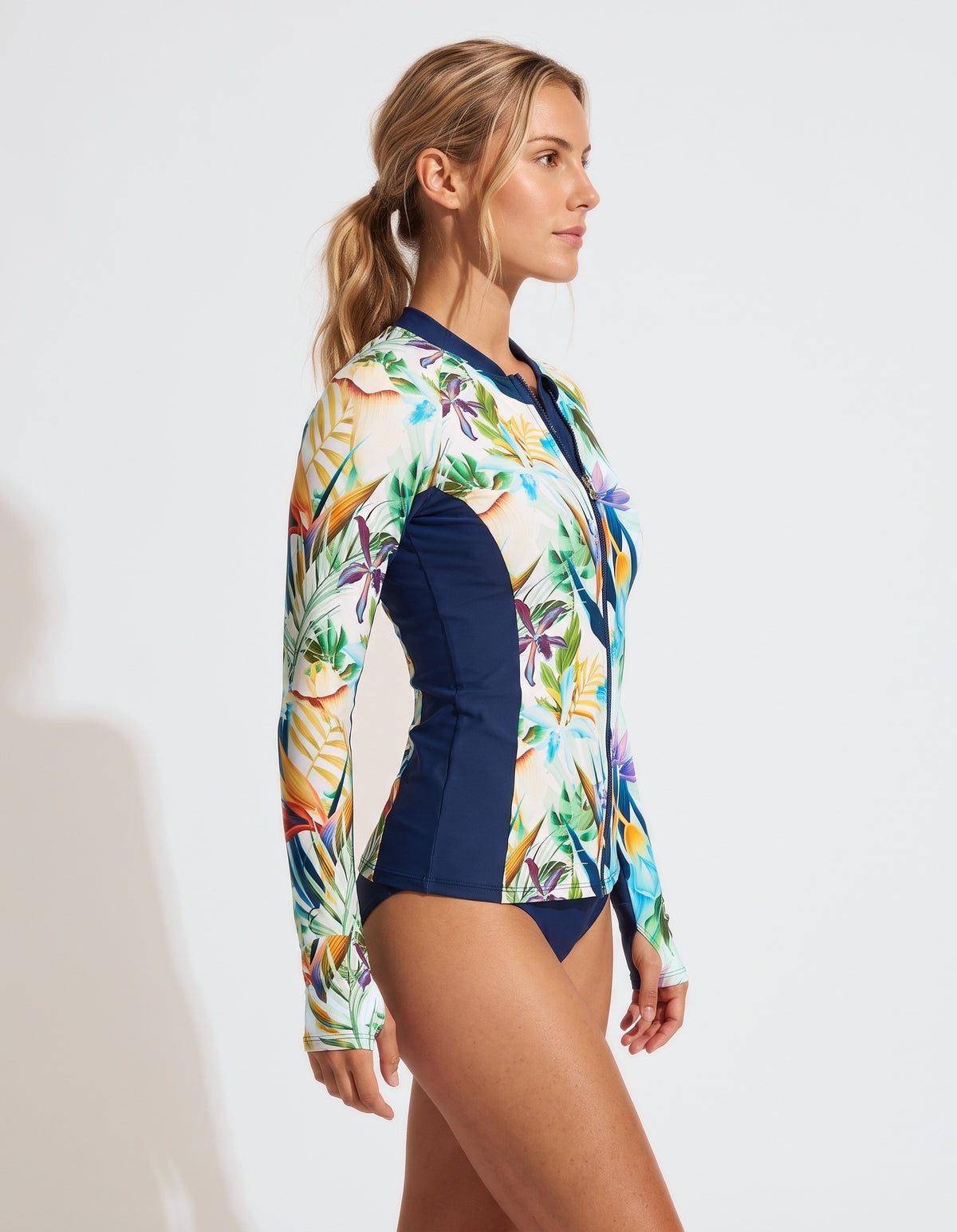 A woman modeling a long-sleeve swim shirt with a tropical floral print on the front and navy blue side panels, shown against a plain white studio background.