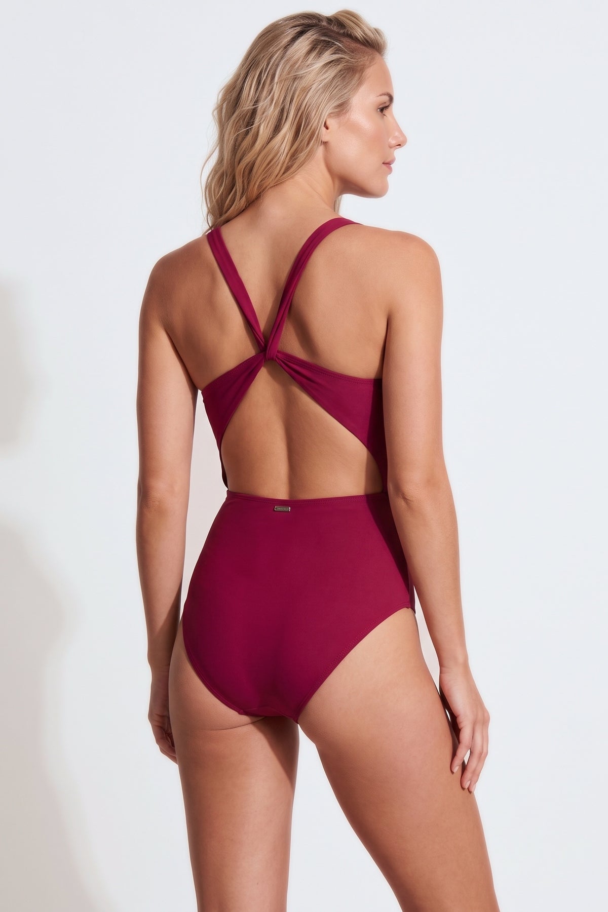 A woman modeling a one-piece swimsuit from the back, showing a crossover crisscross strap design and side cutouts, in a deep burgundy color against a plain white studio background.