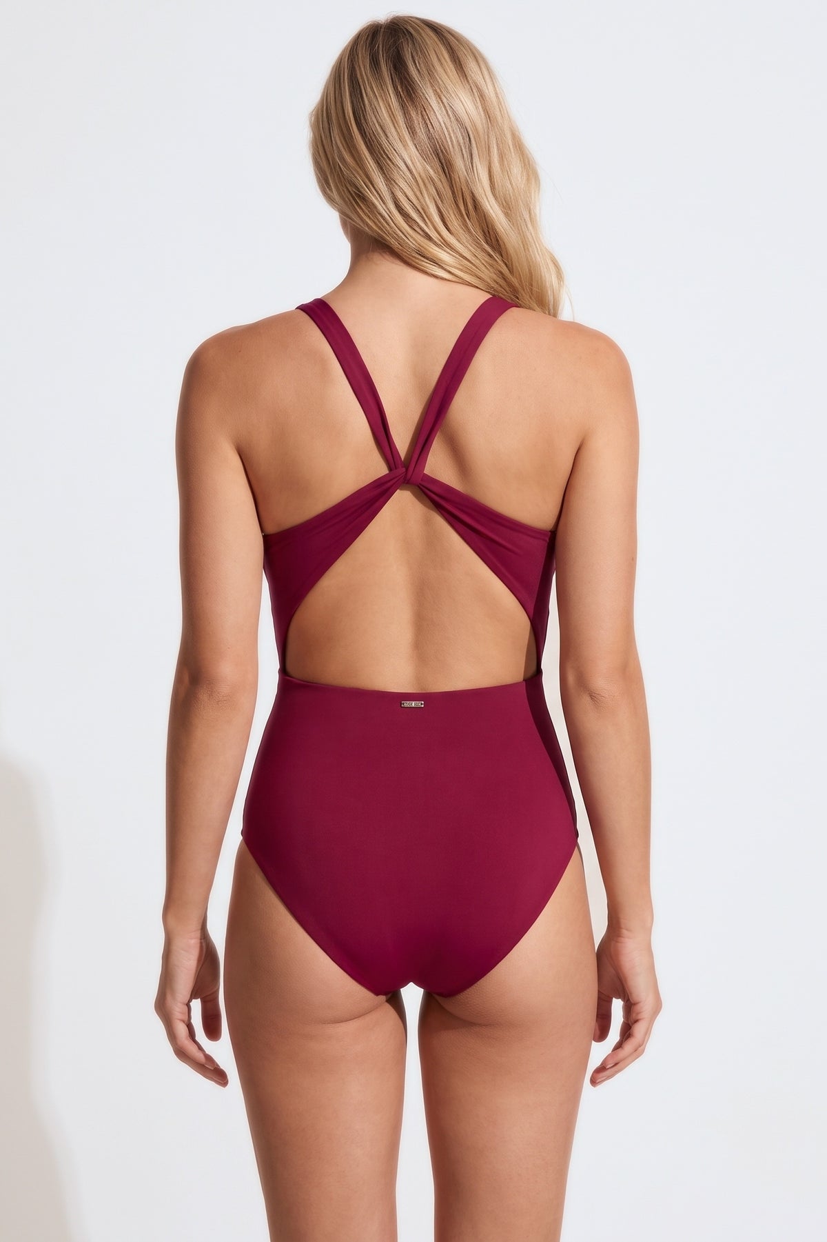 A woman viewed from the back wearing a high-waisted one-piece swimsuit in deep burgundy. The swimsuit features crisscrossing back straps and a small gold logo on the center back waistband.