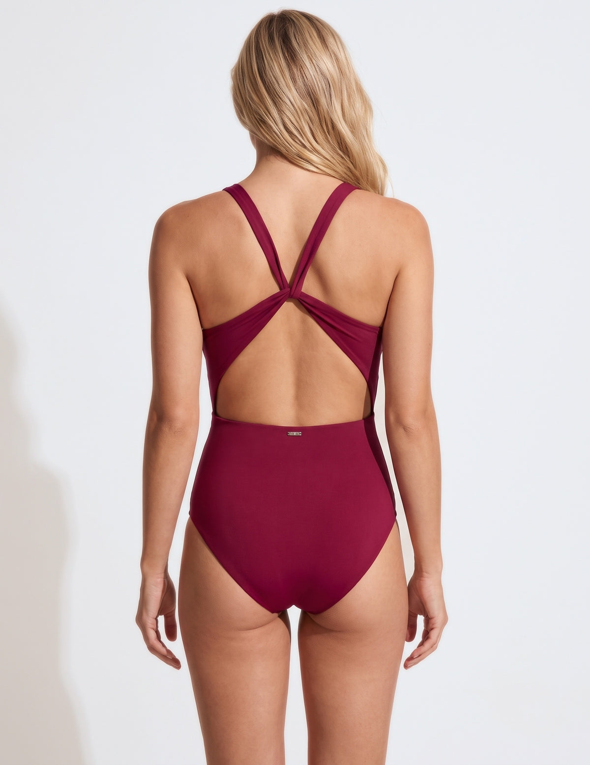 A woman viewed from the back wearing a high-waisted one-piece swimsuit in deep burgundy. The swimsuit features crisscrossing back straps and a small gold logo on the center back waistband.