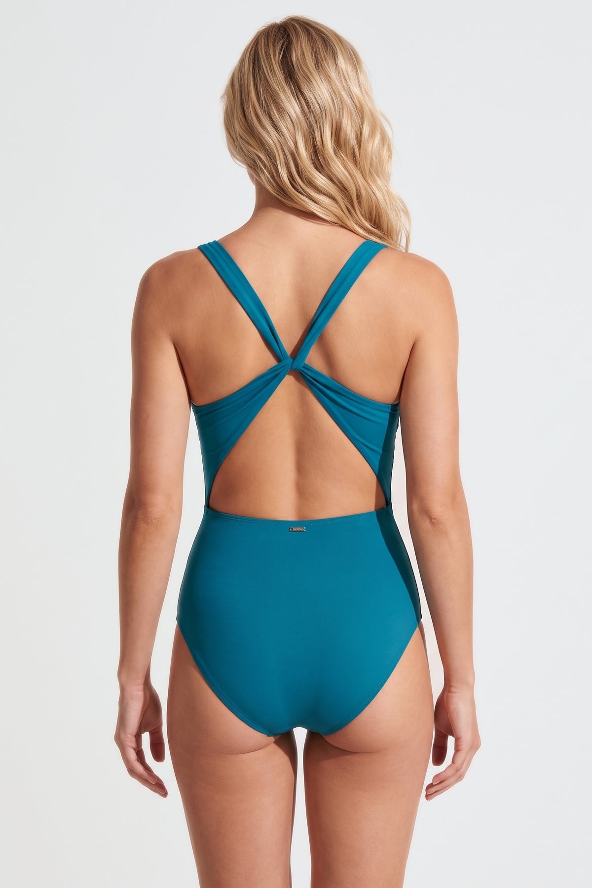 A woman shown from the back wearing a teal one-piece swimsuit with crisscrossing straps across the upper back, photographed against a clean light gray studio background.