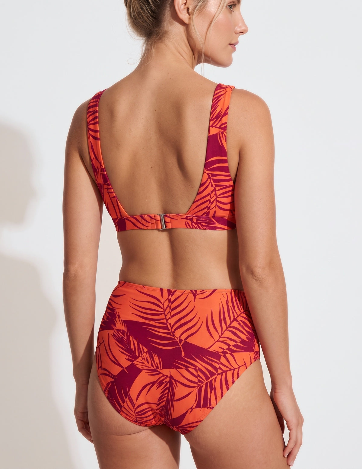 A woman shown from the back wearing a two-piece bikini with a bold tropical leaf print in bright orange and deep magenta-red. The bikini features a low open back on the top and high-waisted bottoms, photographed against a clean white studio background.