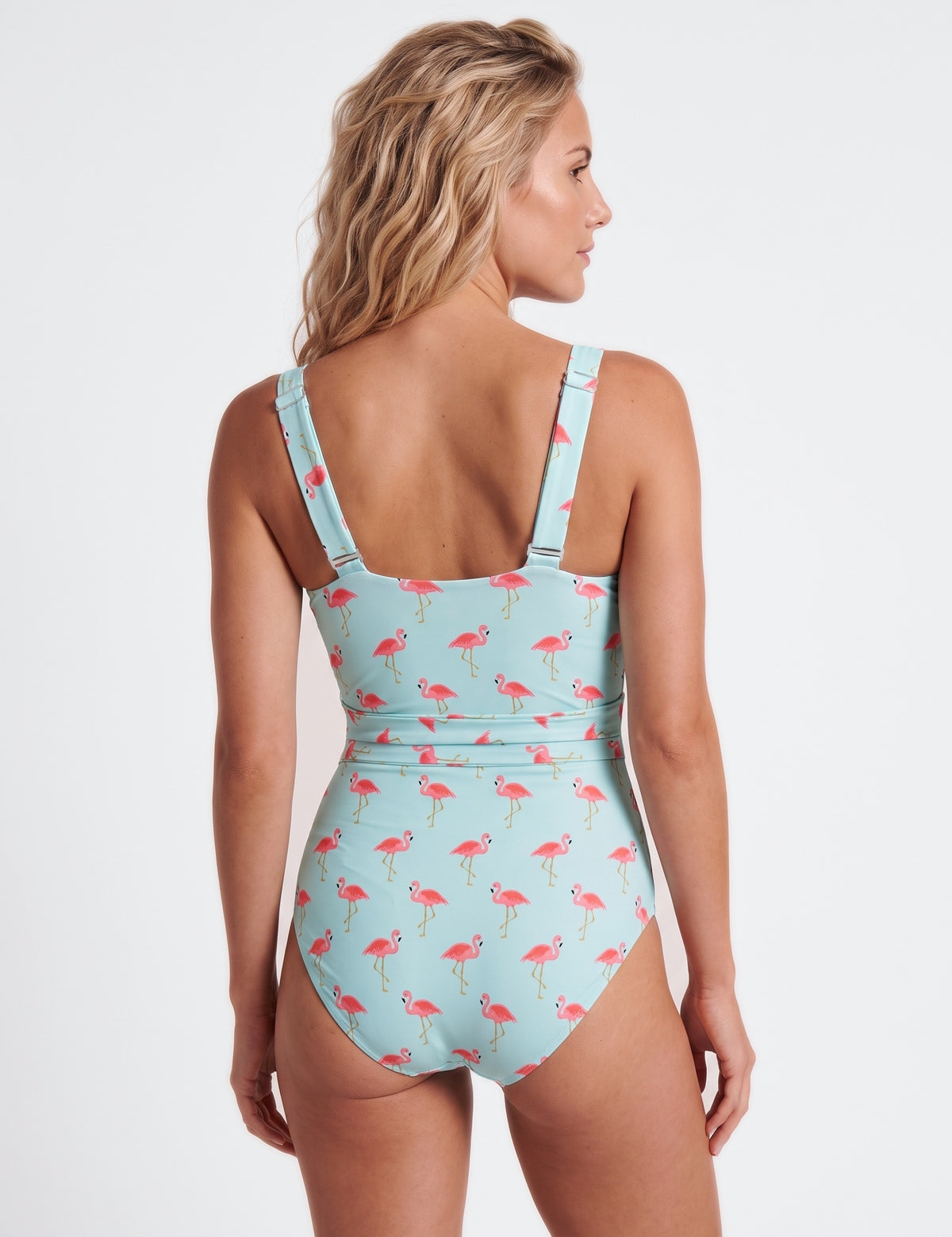 A woman shown from the back wearing a one-piece swimsuit with a light blue background and an allover pink flamingo print, against a plain light gray studio background.