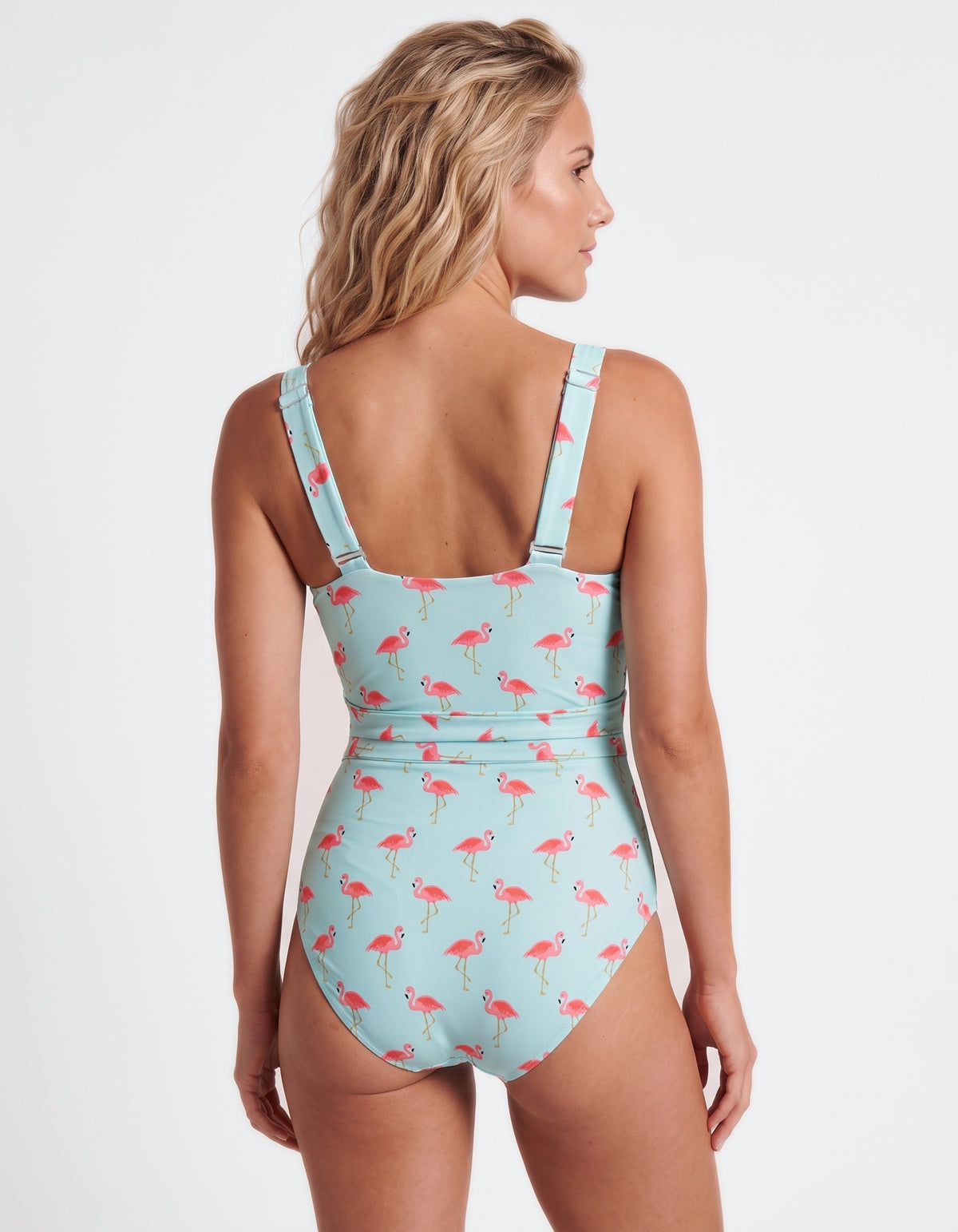 A woman shown from the back wearing a one-piece swimsuit with a light blue background and an allover pink flamingo print, against a plain light gray studio background.