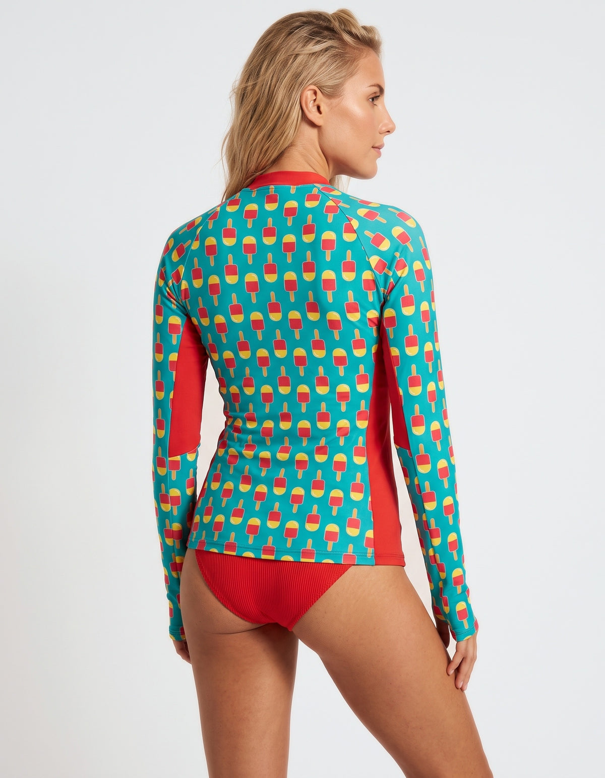 A woman modeling a long-sleeve swim top from the back, featuring a turquoise background with a repeating red and yellow ice cream cone pattern and solid red side panels and bikini bottom.