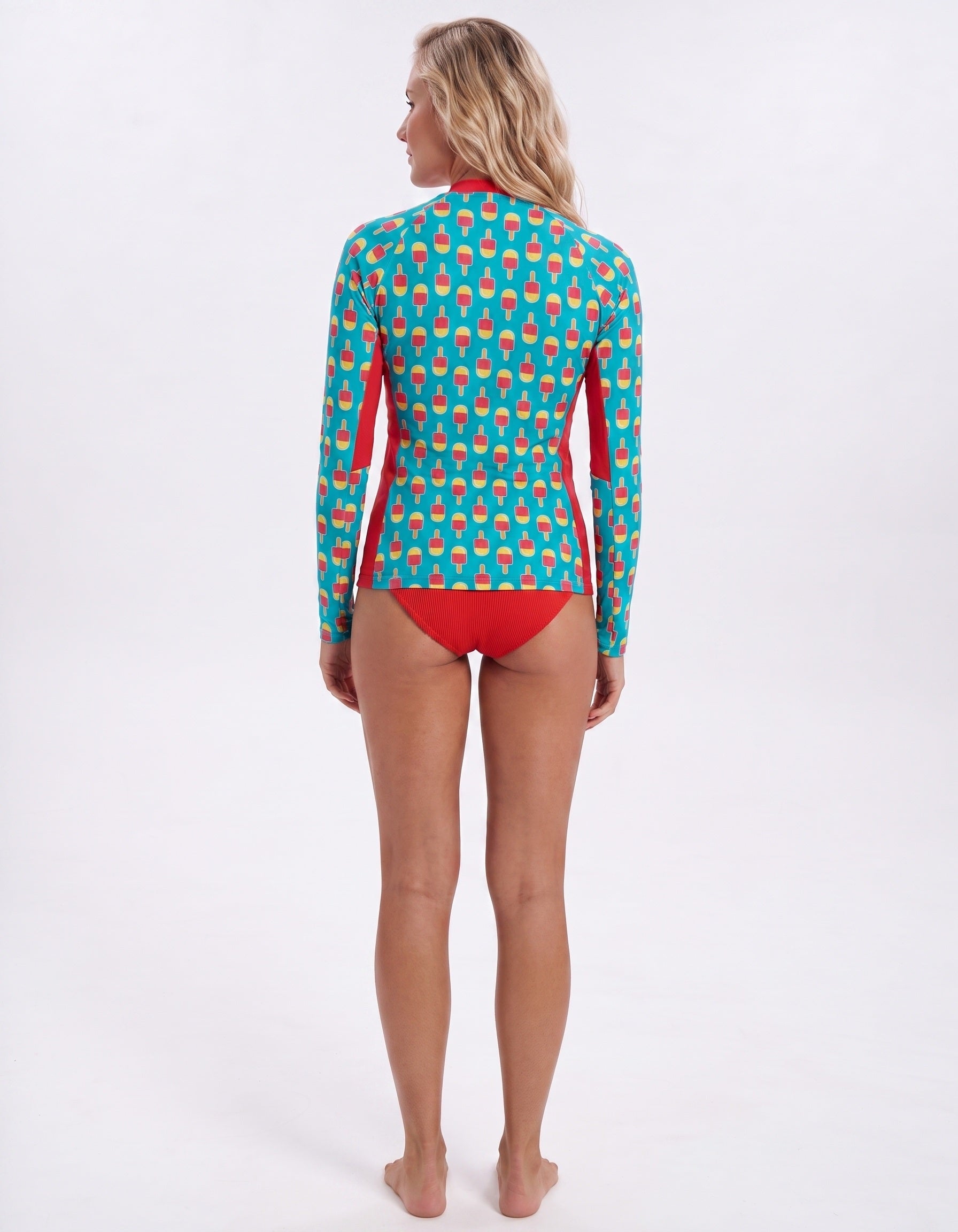 A woman stands barefoot against a plain white studio background, photographed from the back while wearing a long-sleeve swim top with a turquoise and coral geometric pattern and solid red side panels and bikini bottom.