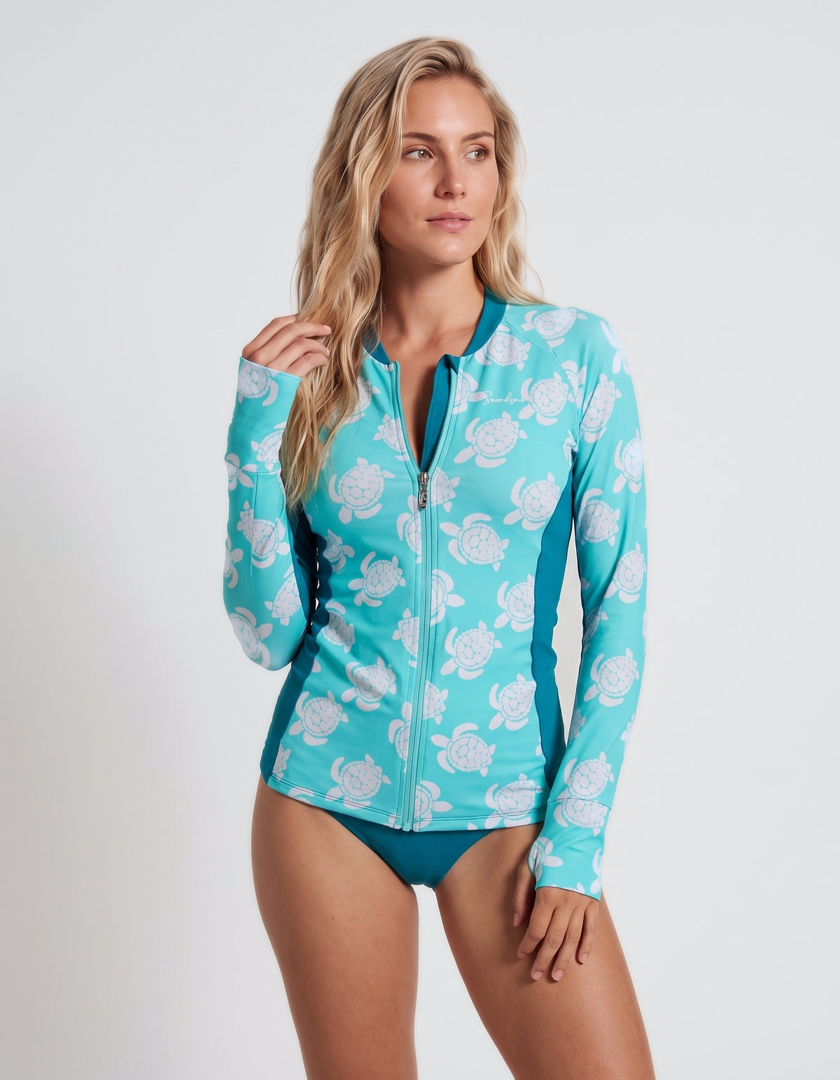 A woman modeling a turquoise and white floral print long-sleeve zip-up swim cover-up with teal side panels, paired with matching teal swim briefs, against a light gray studio background.