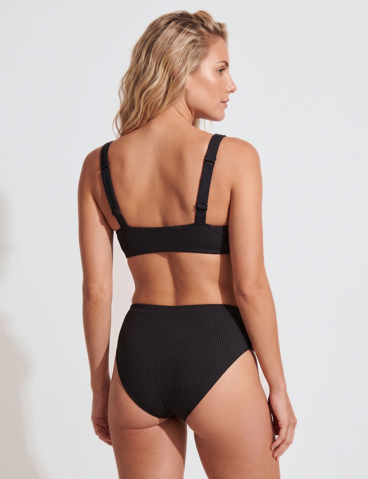 A woman shown from the back wearing a black ribbed bikini set against a plain light gray studio background.