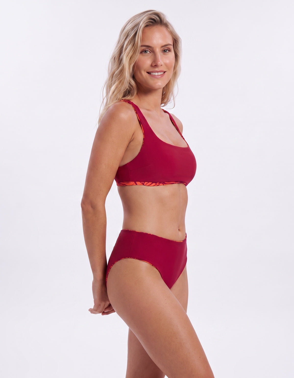 A woman modeling a two-piece bikini set in a deep red color, featuring a sports bra top with a subtle orange patterned trim and matching high-waisted bottoms, set against a plain white studio background.