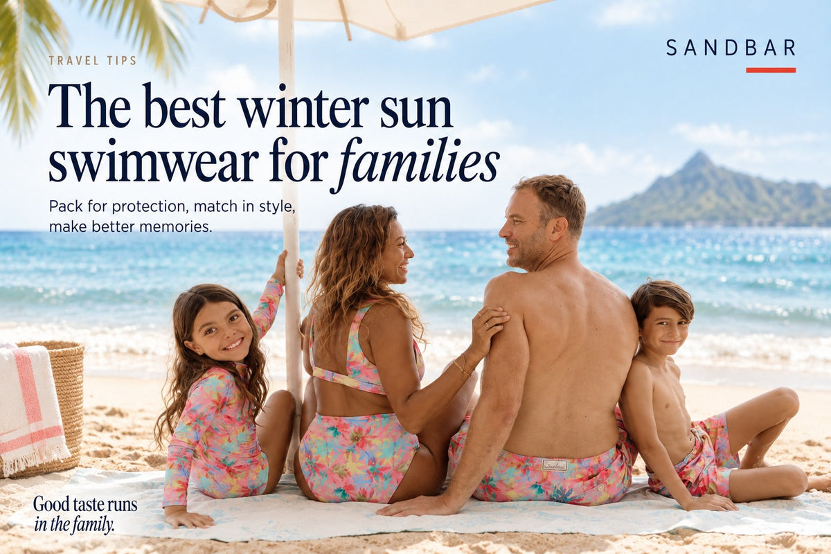 A family of four relaxing on a tropical beach in matching Sandbar Pink Palm swimwear, with turquoise waters and lush mountains behind them. The image features the headline “The best winter sun swimwear for families.