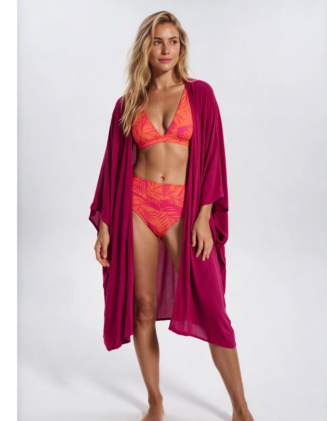 A woman modeling a two-piece bikini in bright orange and pink with a matching magenta open-front cover-up on a light gray studio background. Sandbar Swimwear