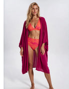 A woman modeling a two-piece bikini in bright orange and pink with a matching magenta open-front cover-up on a light gray studio background. Sandbar Swimwear