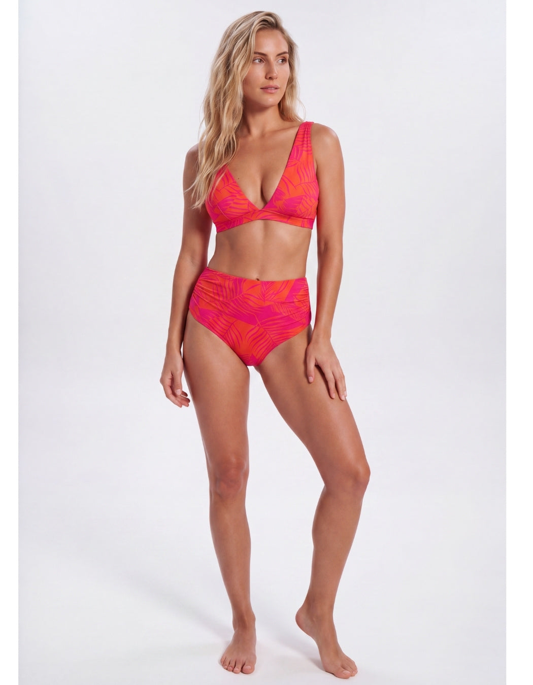 A woman modeling a two-piece bikini swimsuit with a vibrant pink and orange abstract pattern, set against a plain light gray studio background. Sandbar Swimwear