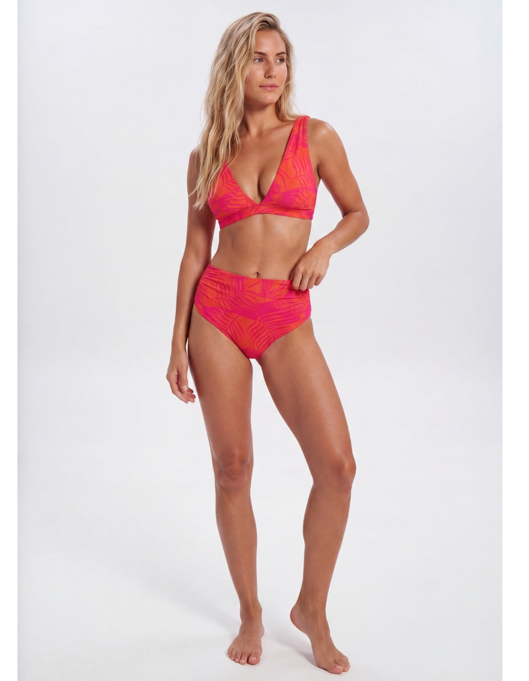 A woman modeling a two-piece bikini swimsuit with a bright pink and orange abstract pattern against a plain light gray studio background. Sandbar Swimwear