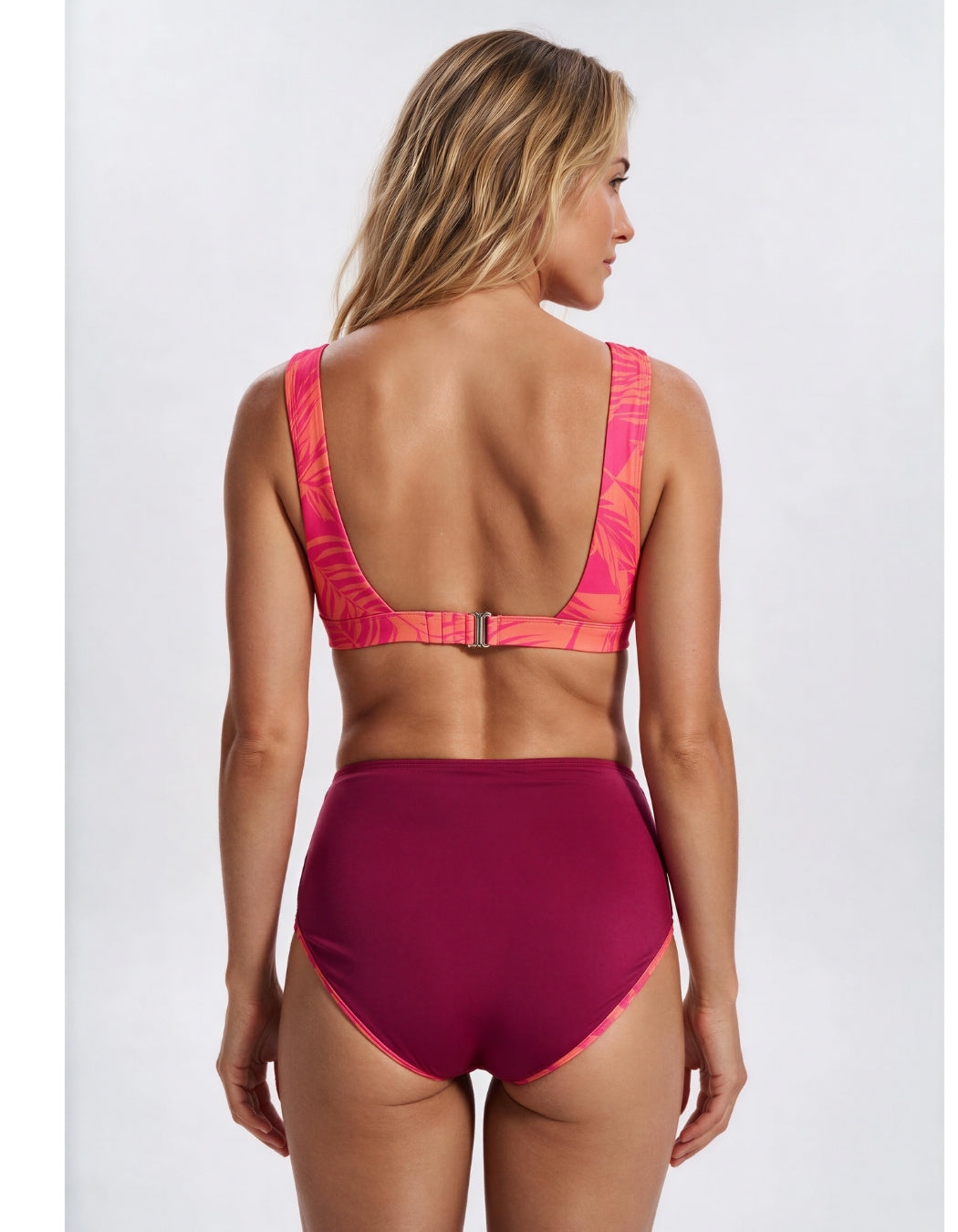A woman shown from the back wearing a two-piece swimsuit with a bright pink and orange abstract patterned bra top and high-waisted magenta bottoms, against a light gray studio background. Sandbar Swimwear