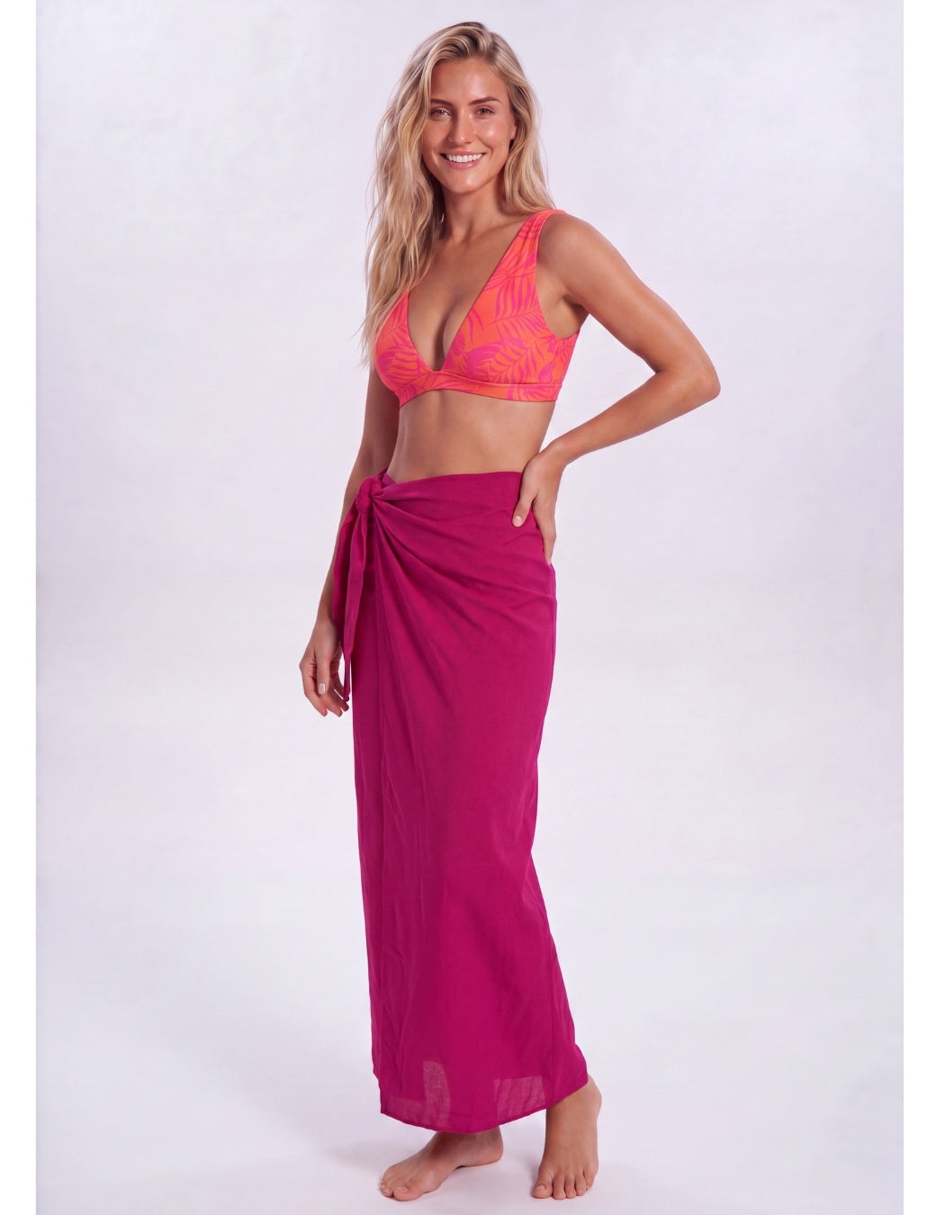 A woman modeling a bright pink patterned bikini top paired with a matching magenta sarong wrap skirt, standing against a light gray studio background. Sandbar Swimwear