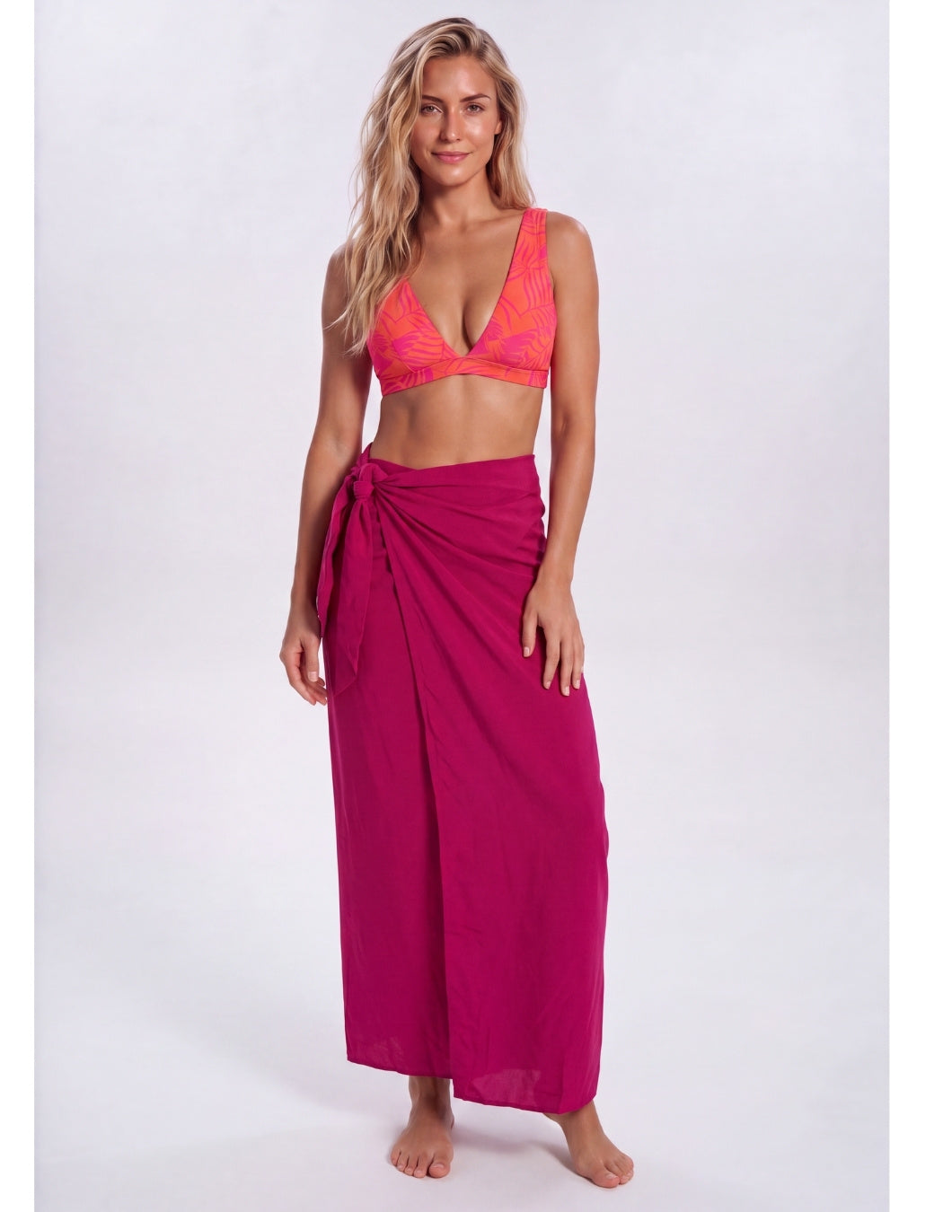 A woman modeling a bright magenta wrap sarong and a matching pink-orange patterned bikini top against a light gray studio background. Sandbar Swimwear