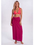 A woman modeling a bright magenta wrap sarong and a matching pink-orange patterned bikini top against a light gray studio background. Sandbar Swimwear