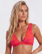 A woman wearing a vibrant bikini top with a pink and orange tropical leaf pattern, photographed against a clean light gray studio background. Sandbar Swimwear