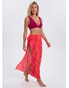 A woman modeling a bright pink and orange tropical leaf-patterned sarong wrap skirt with a matching pink bikini top, standing against a light gray studio background. Sandbar Swimwear